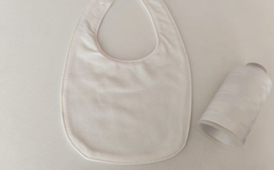Fotografia di un bavaglino bianco per neonati messo su un tavolo bianco con accanto un filo da ricamo bianco.