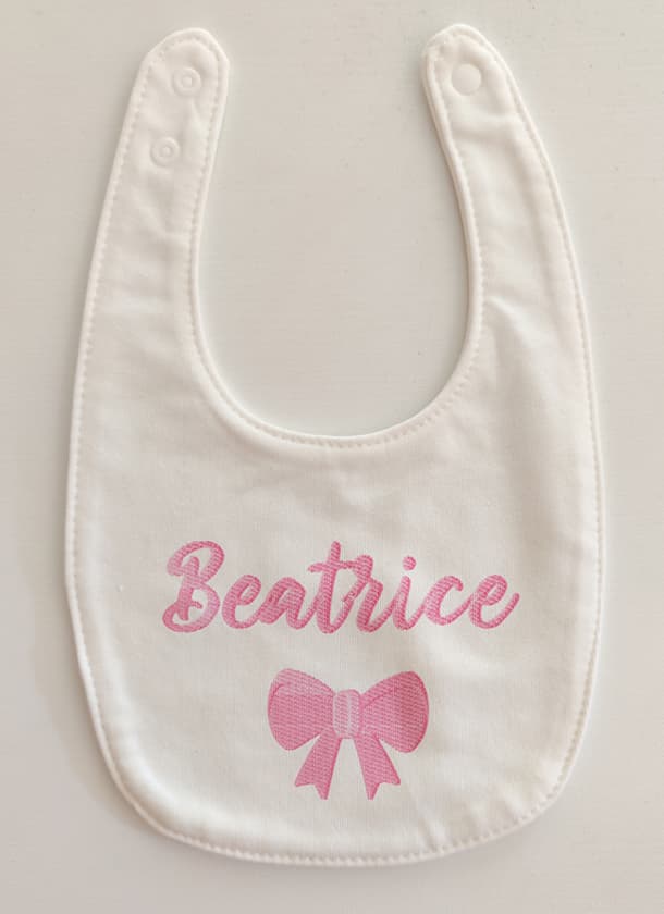Fotografia di un bavaglino per neonato bianco con ricamato in rosa il nome "Beatrice" ed un fiocco.