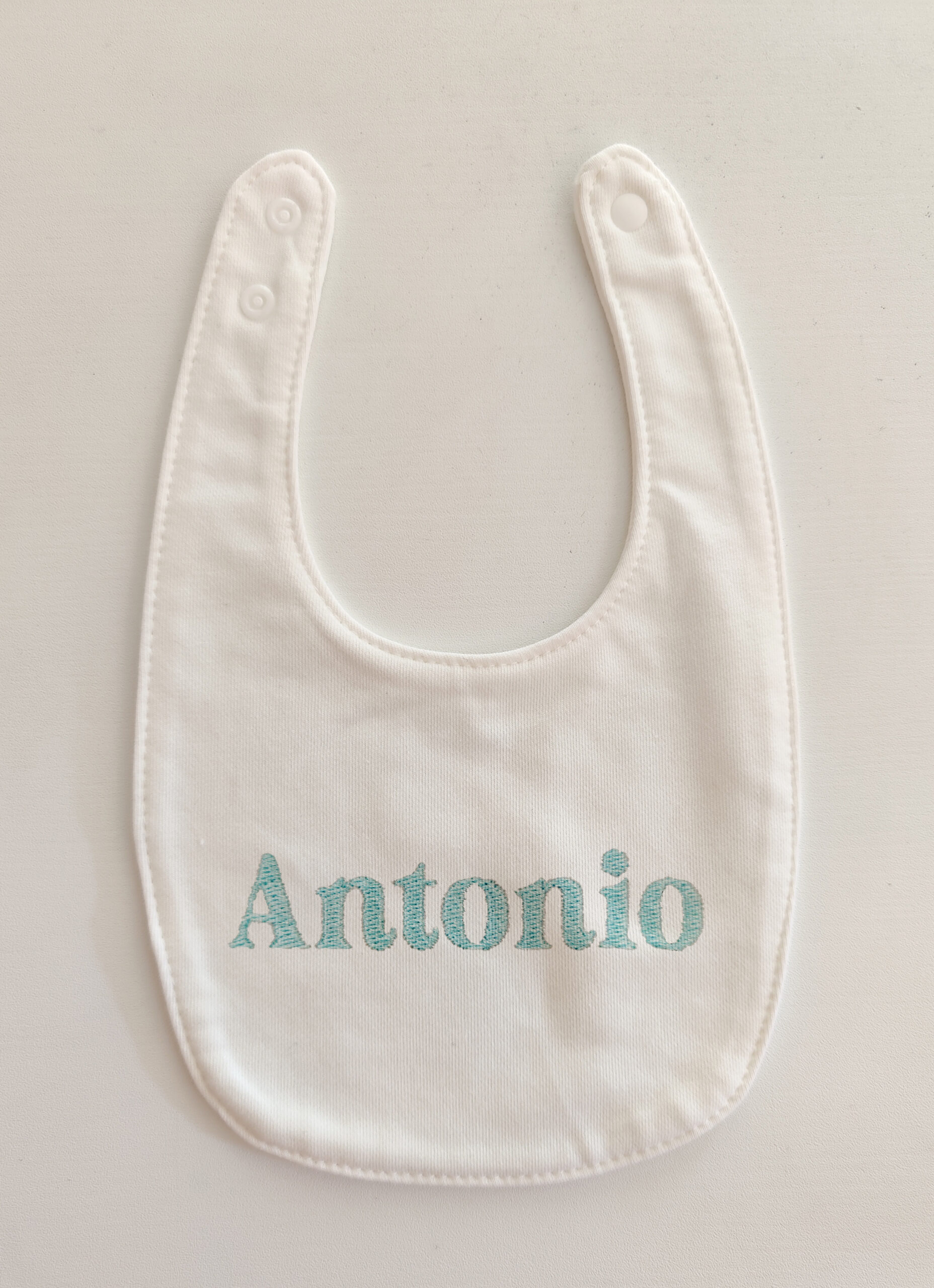 Fotografia di un bavaglino per neonato bianco con ricamato il nome "Antonio" in azzurro.