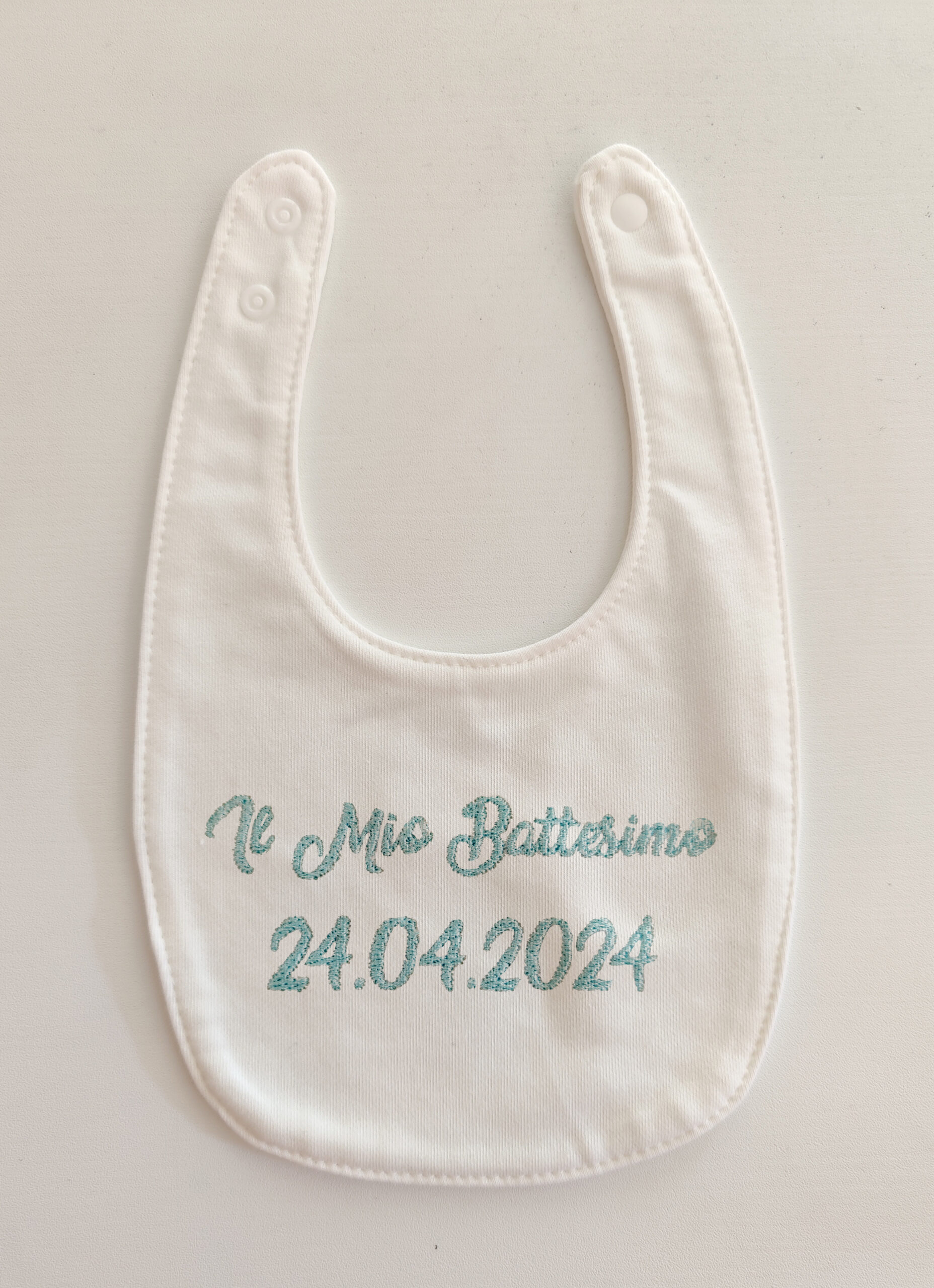 Fotografia di un bavaglino bianco con ricamato in azzurro "Il mio battesimo" e la data.
