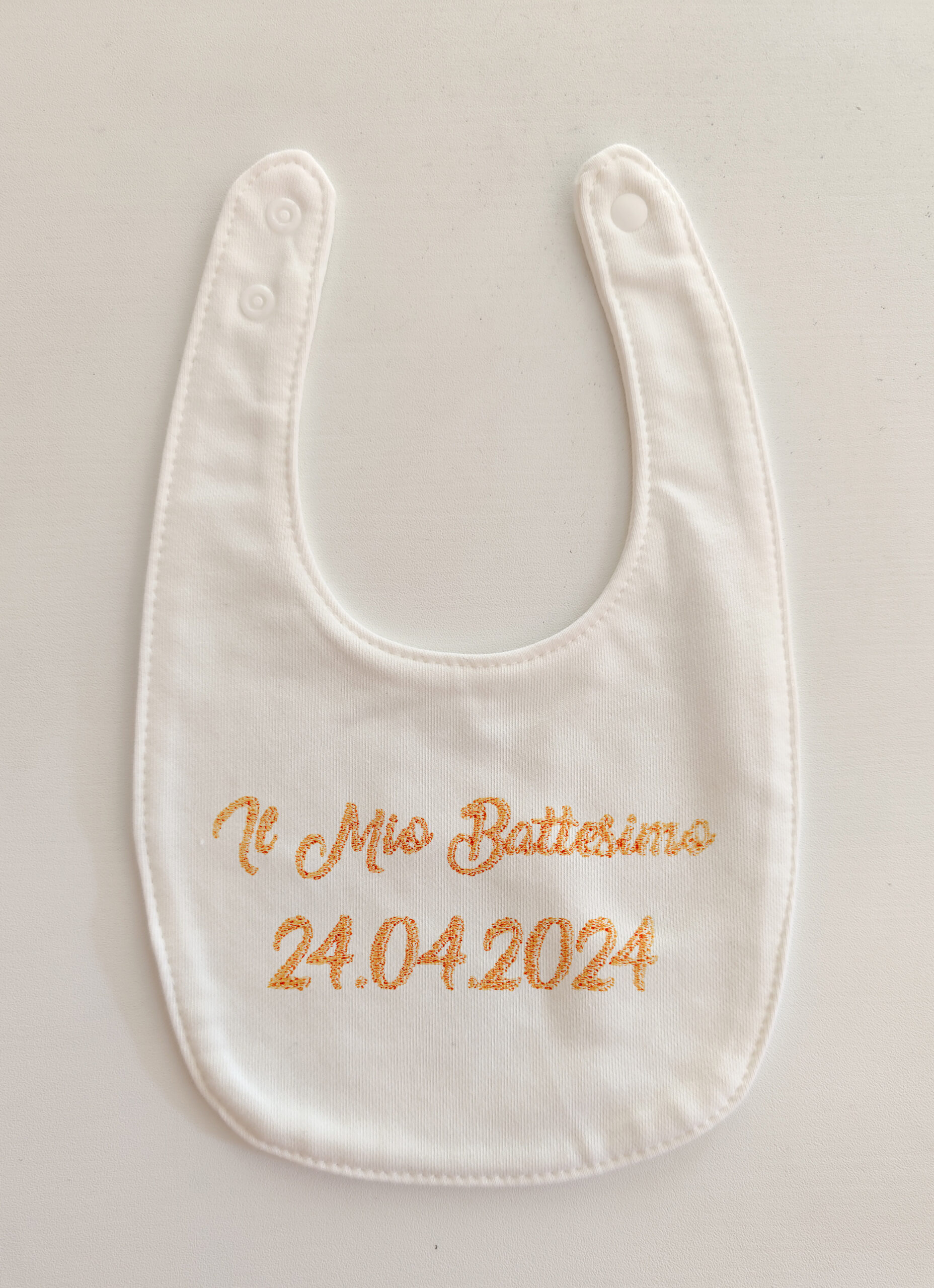 Fotografia di un bavaglino bianco con ricamato in rosa "Il mio battesimo" e la data.