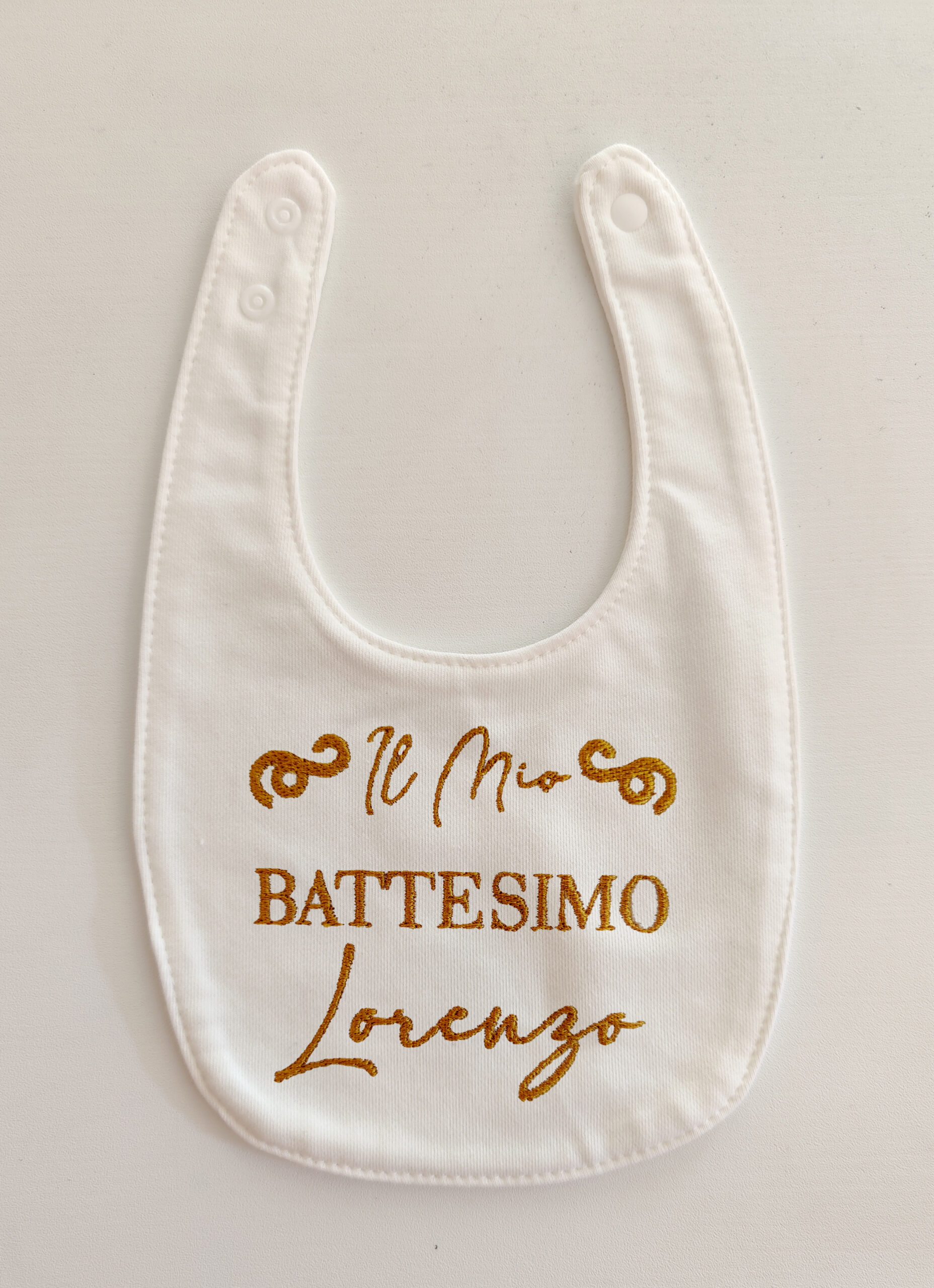 Foto di un bavaglino per neonati bianco con ricamato in oro la scritta "Il Mio Battesimo Lorenzo"
