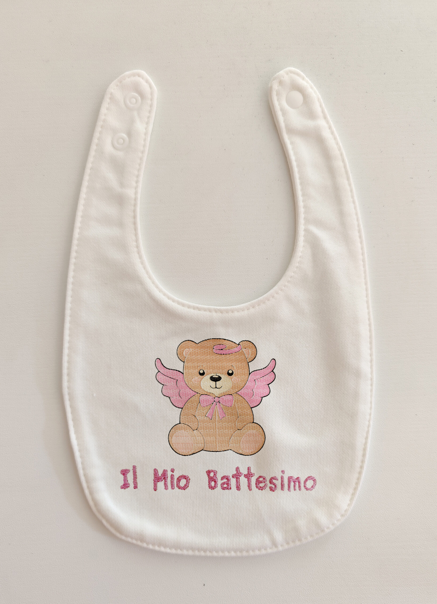 Fotografia di un bavaglino per neonati bianco con il ricamo rosa di un orsetto con le ali e la scritta sotto in rosa "Il mio Battesimo".
