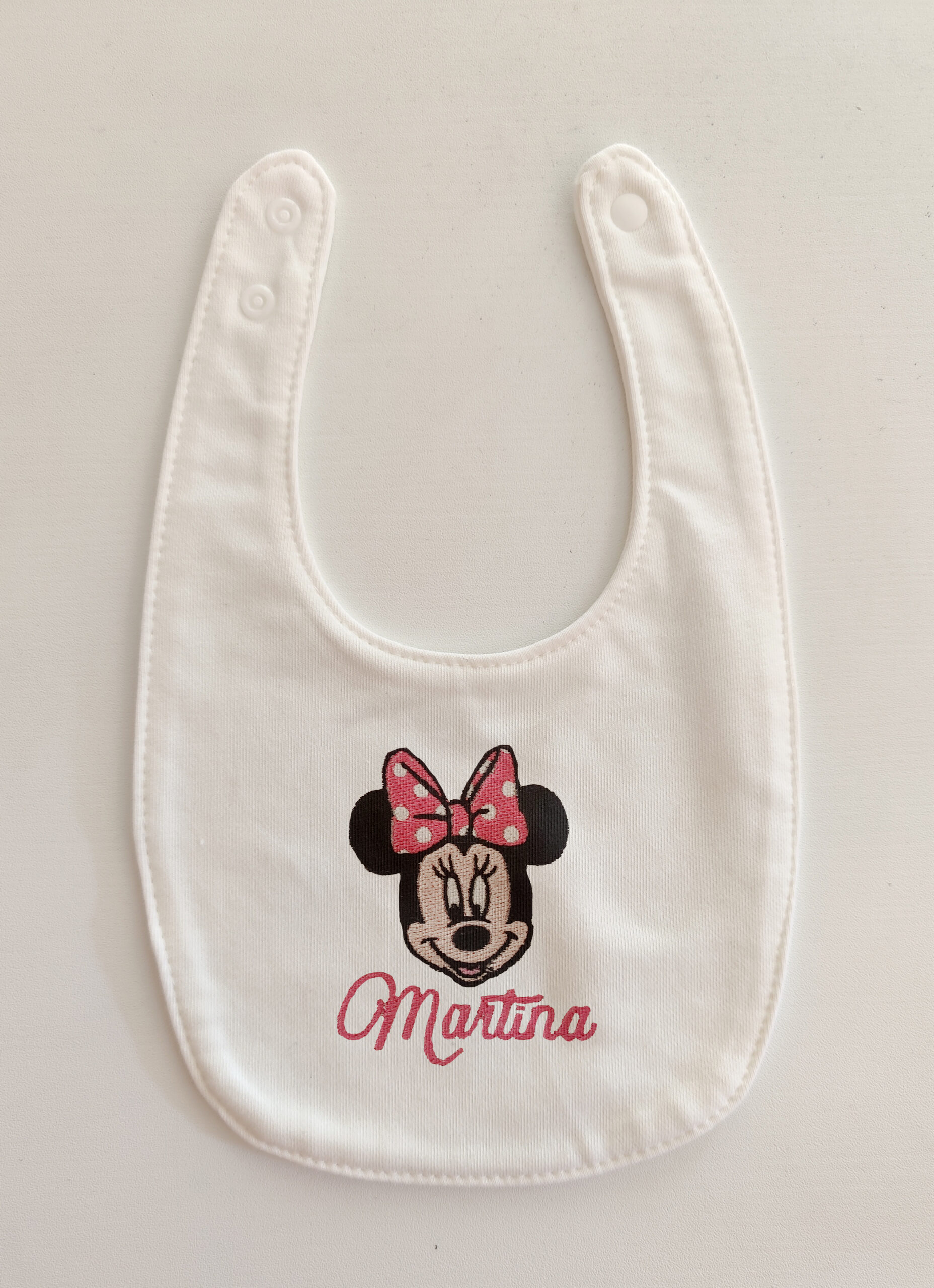 Fotografia di un bavaglino per neonati bianco con sopra ricamata la testa di Minnie Mouse ed il nome Martina in rosa.