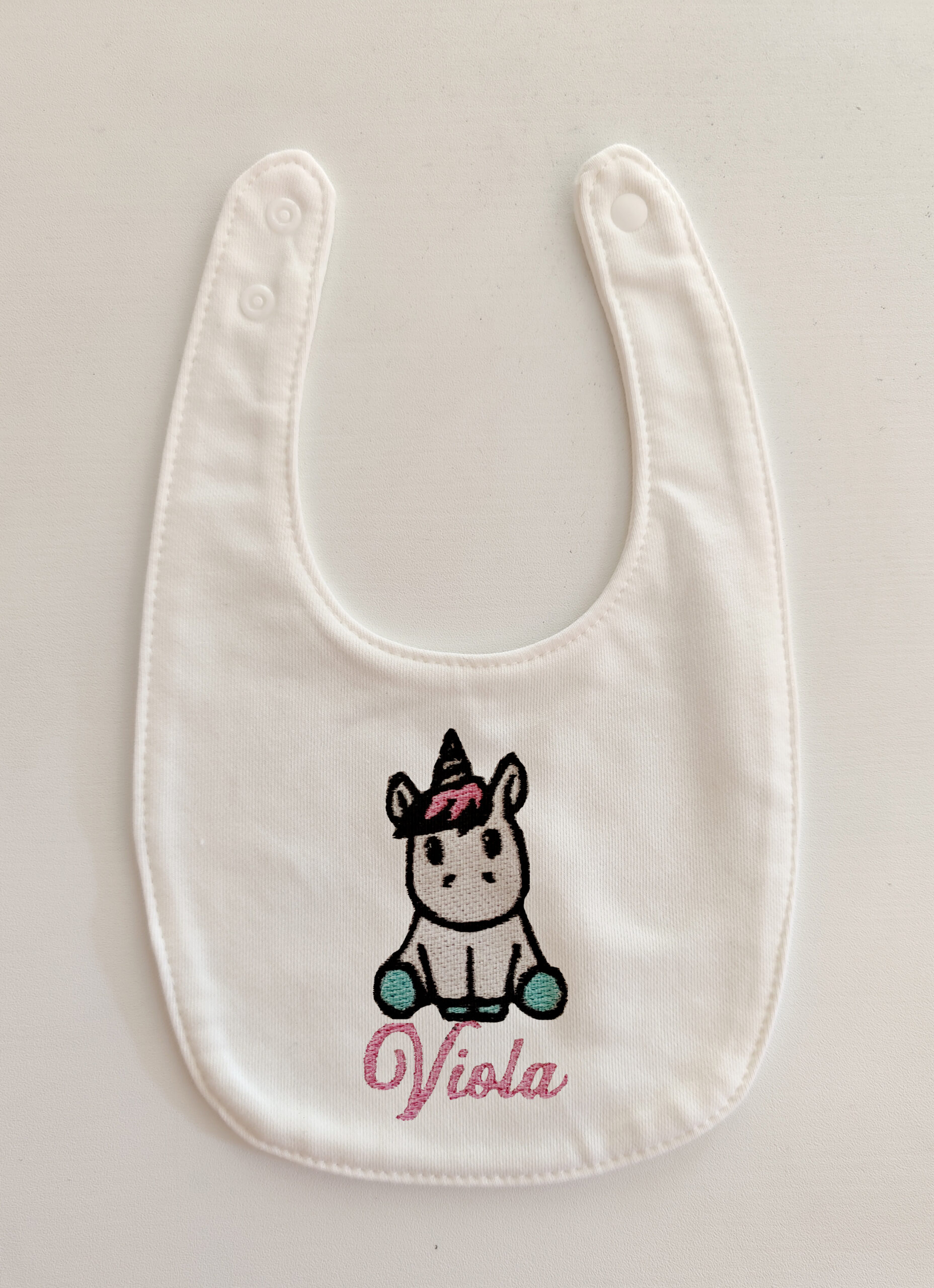 Fotografia di un bavaglino per neonati bianco con ricamato un unicorno seduto ed il nome "Viola" in rosa.