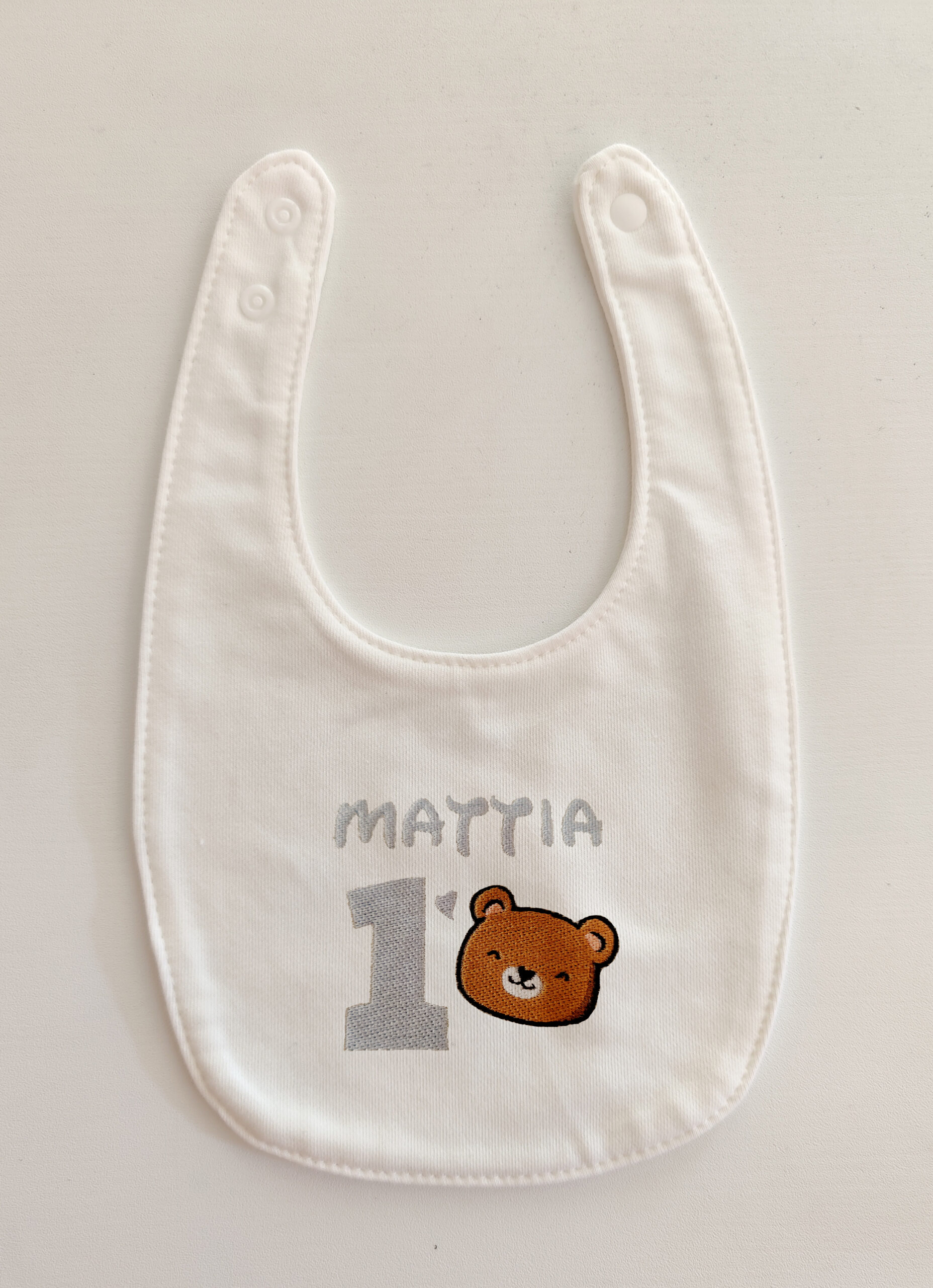 Fotografia di un bavaglino per neonati bianco con ricamato la testa di un orsetto ed il nome "Mattia" ed il numero "1" in azzurro.