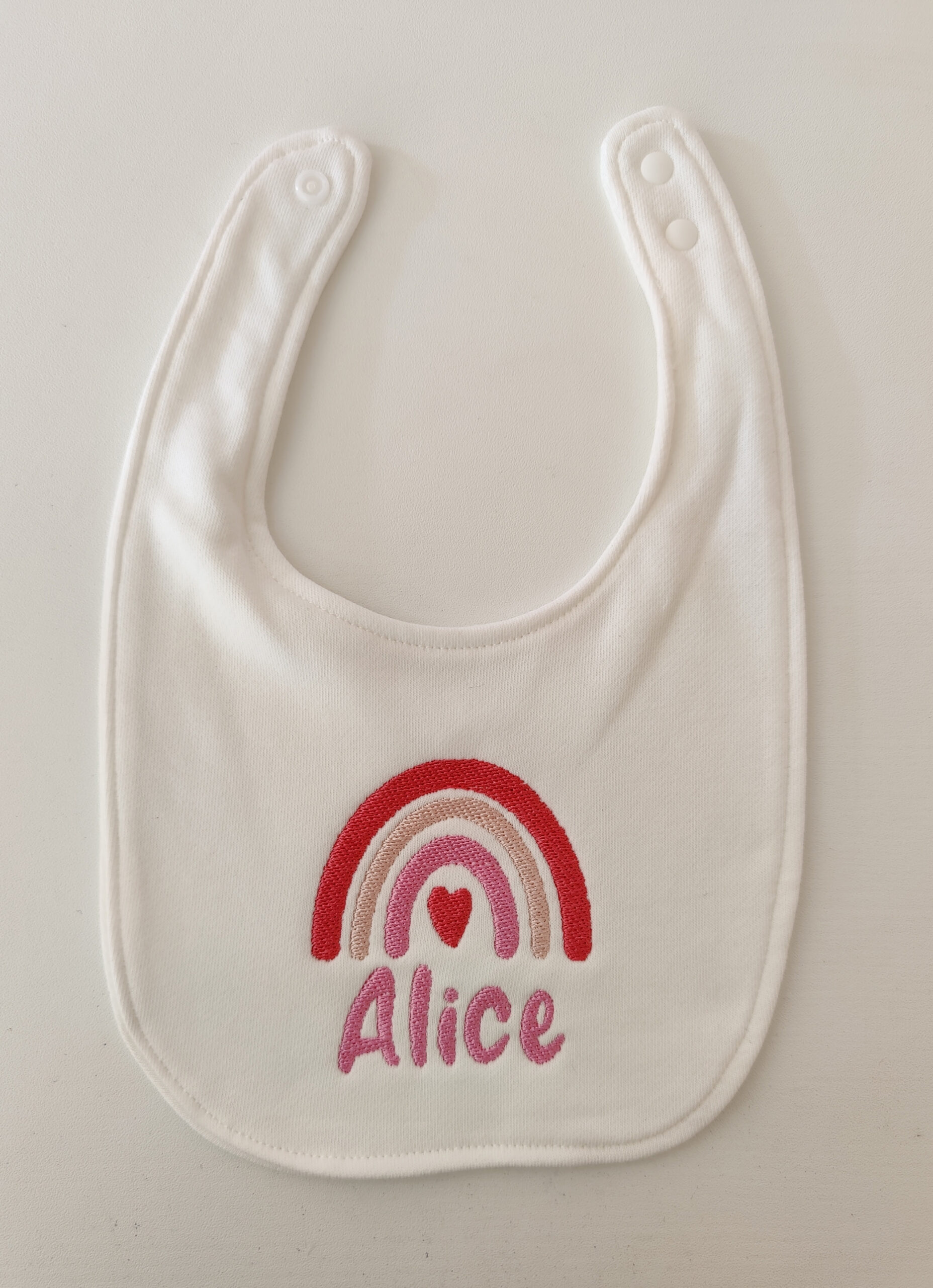 Fotografia di un bavaglino per neonato con ricamato un arcobaleno dalle sfumature rosa ed il nome "Alice" in rosa.