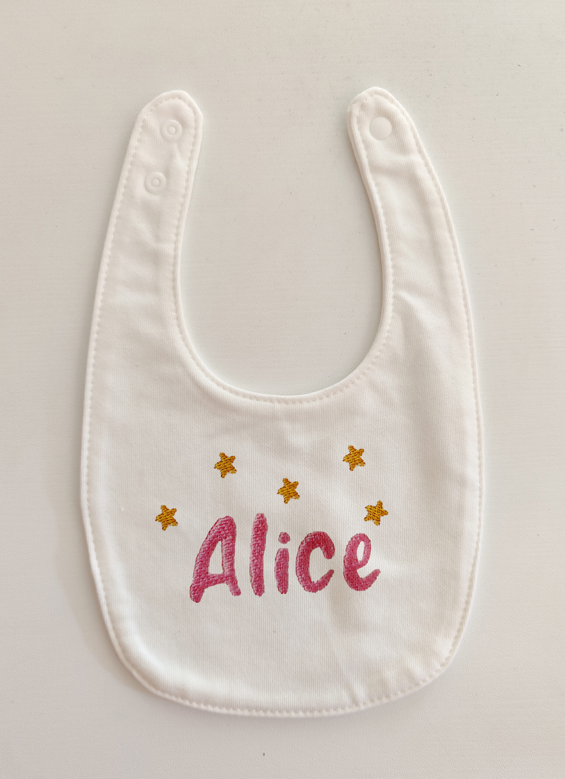 Foto di un bavaglino per neonati bianco con ricamato il nome Alice in rosa e cinque stelle in giallo.