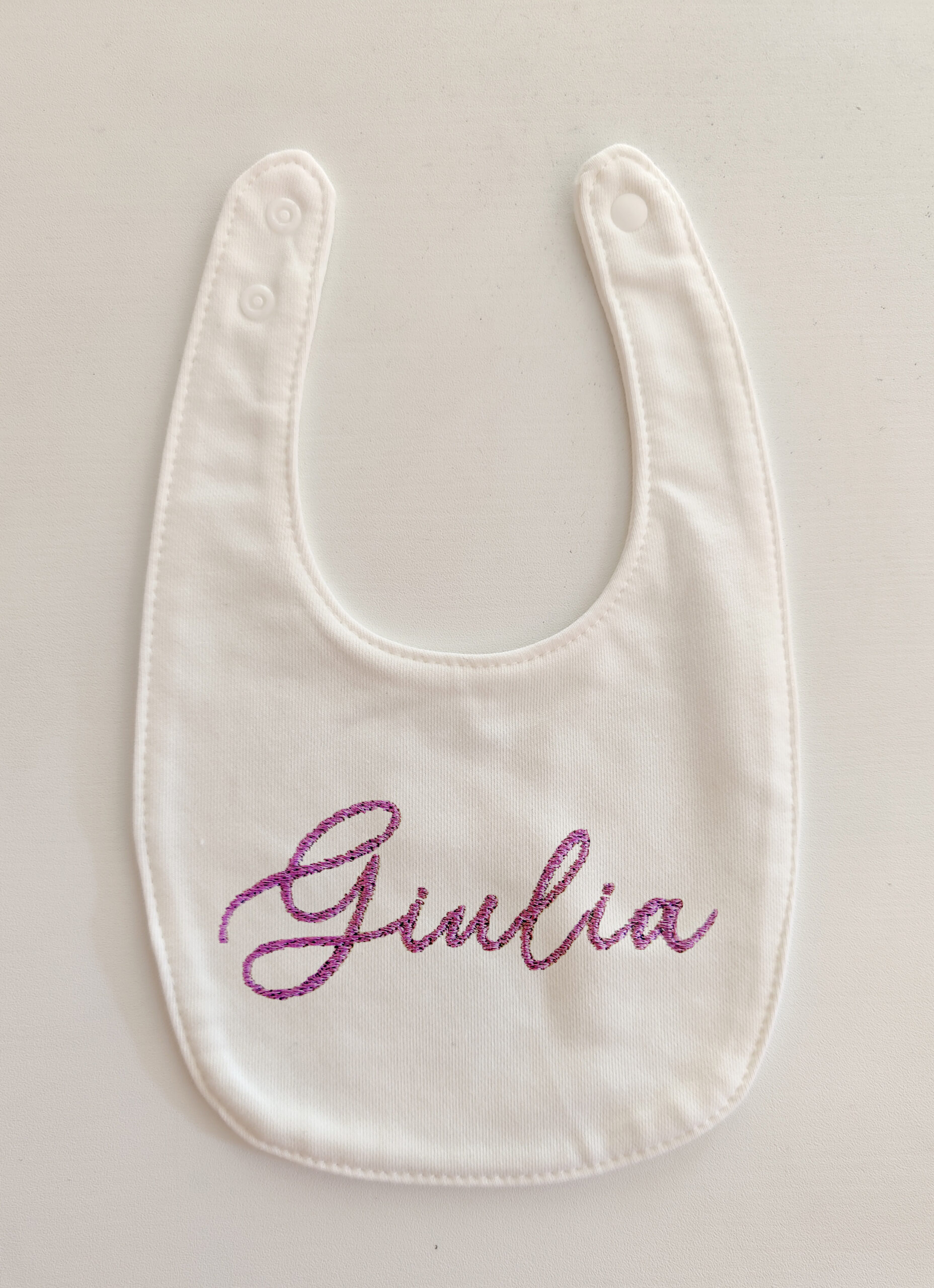 Fotografia di un bavaglino per neonato bianco con ricamato il nome "Giulia" in viola.