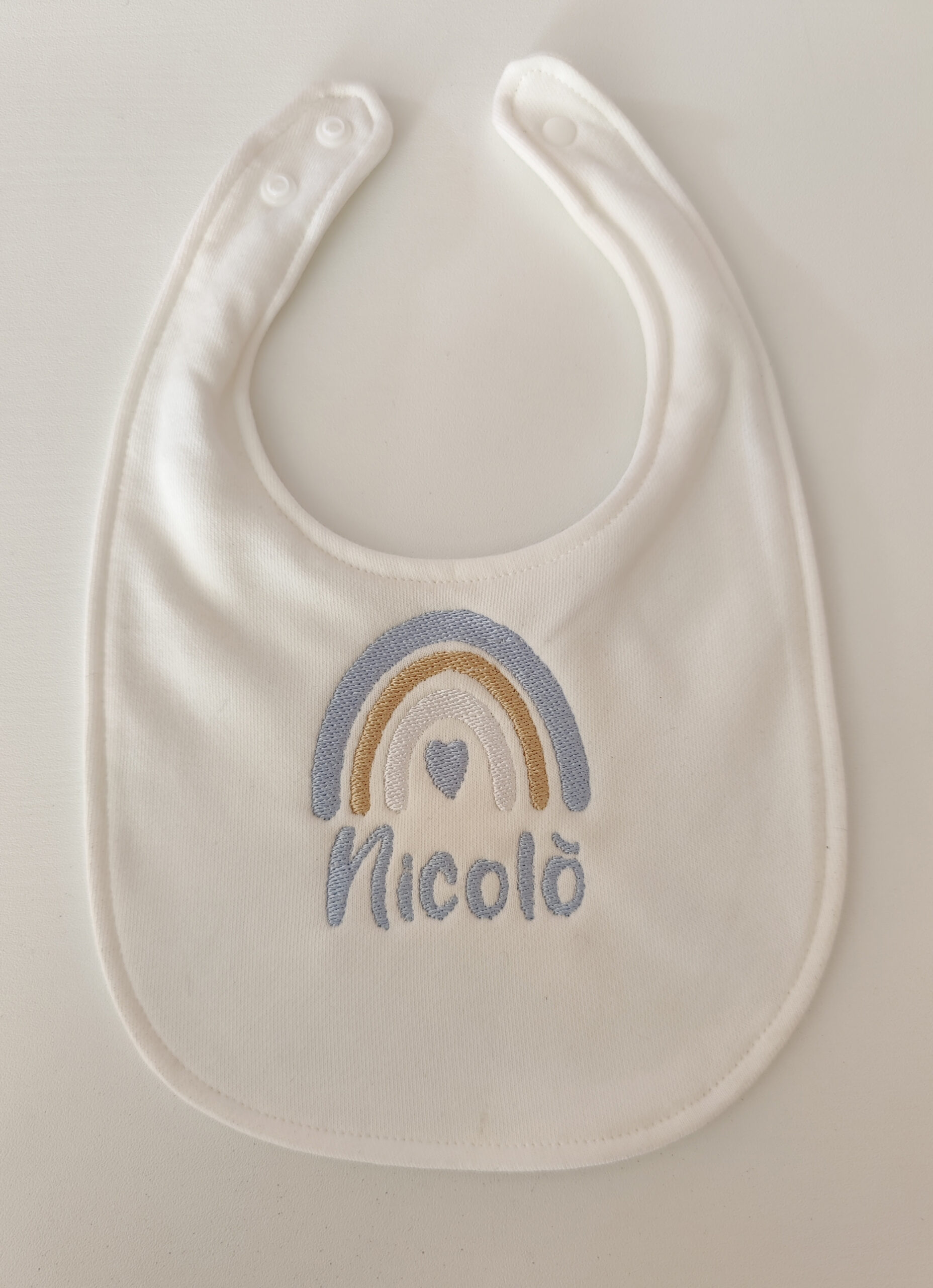 Fotografia di un bavaglino bianco per neonato con ricamato il nome e l'arcobaleno con tonalità pastello.