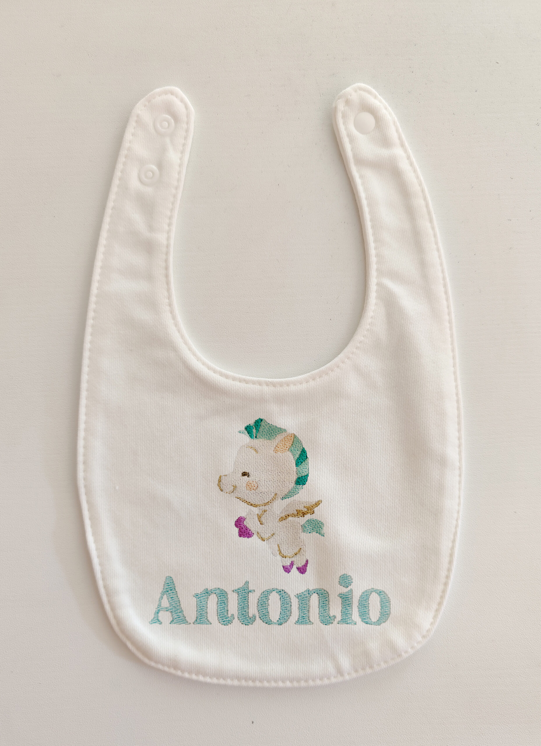 Fotografia di un bavaglino per neonati bianco con ricamato Baby Pegaso del cartone disney "Hercules" e sotto il nome "Antonio" in azzurro.