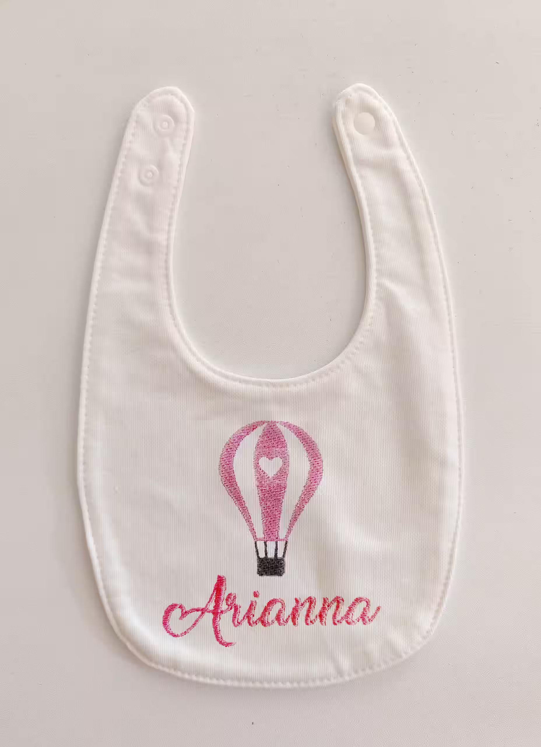 Fotografia di un bavaglino per neonati bianco con ricamo di una mongolfiera rosa e con il nome "Arianna" ricamato in rosa.