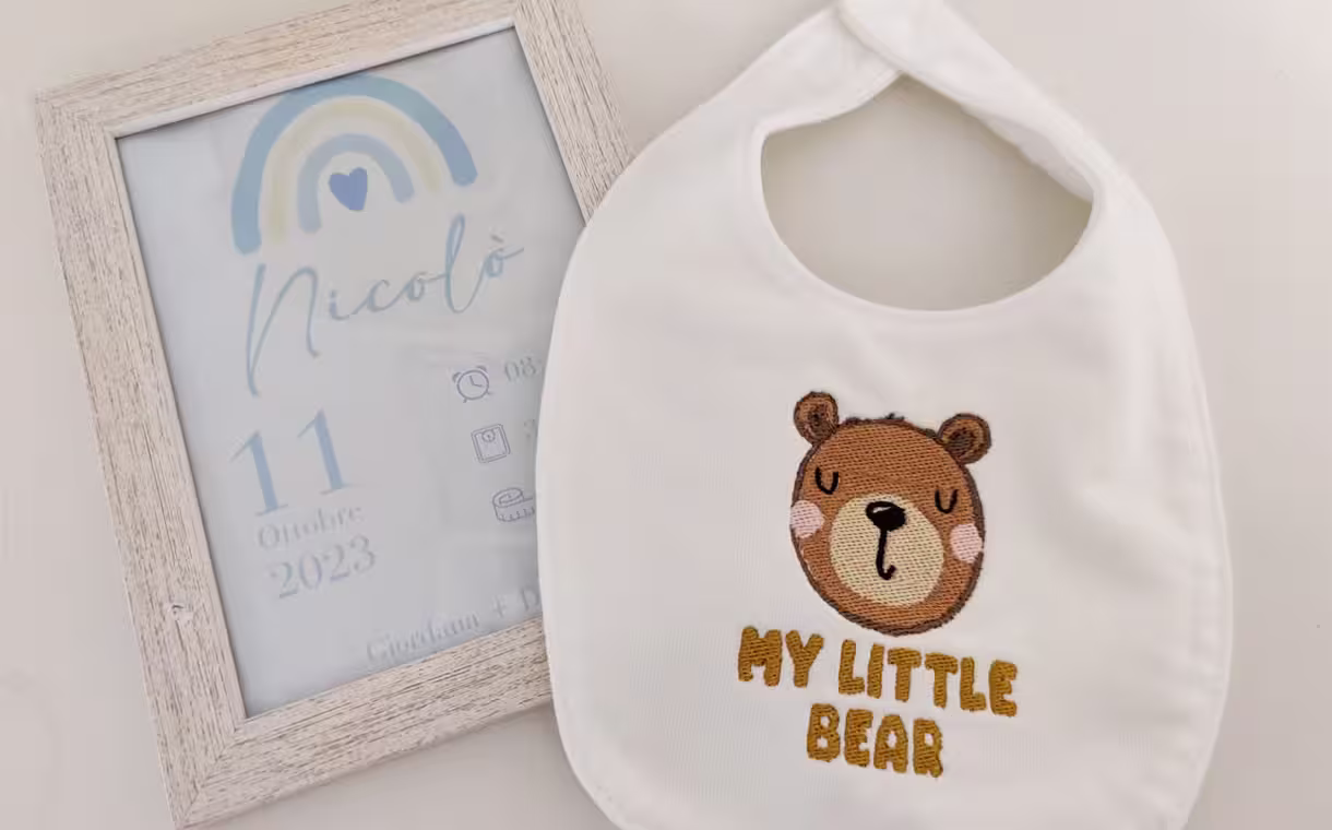 Fotografia di un bavaglino per neonati ricamato con scritto "My Little Bear" e la faccia di un orso sopra un quadretto con i dati nascita di un bambino.