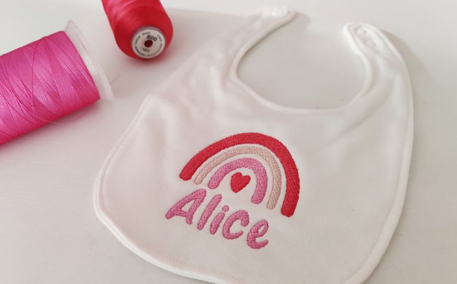 Fotografia di un bavaglino per neonato con ricamato sopra il nome Alice in rosa ed un arcobaleno con le tonalità del rosa. In alto a sinistra ci sono due filati, uno rosa ed uno rosso.