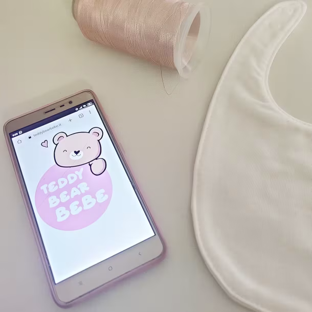 Fotografia di uno smartphone che mostra immagine logo sito web Teddybearbebe con accanto un bavaglino per neonato ed un filo da ricamo in alto