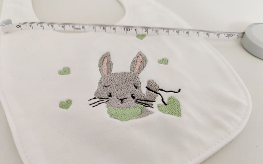 Fotografia in primo piano di un bavaglino bianco con sopra ricamato un coniglio con un cuore a pendolo verde e dei cuoricini intorno. In alto si vede un metro.
